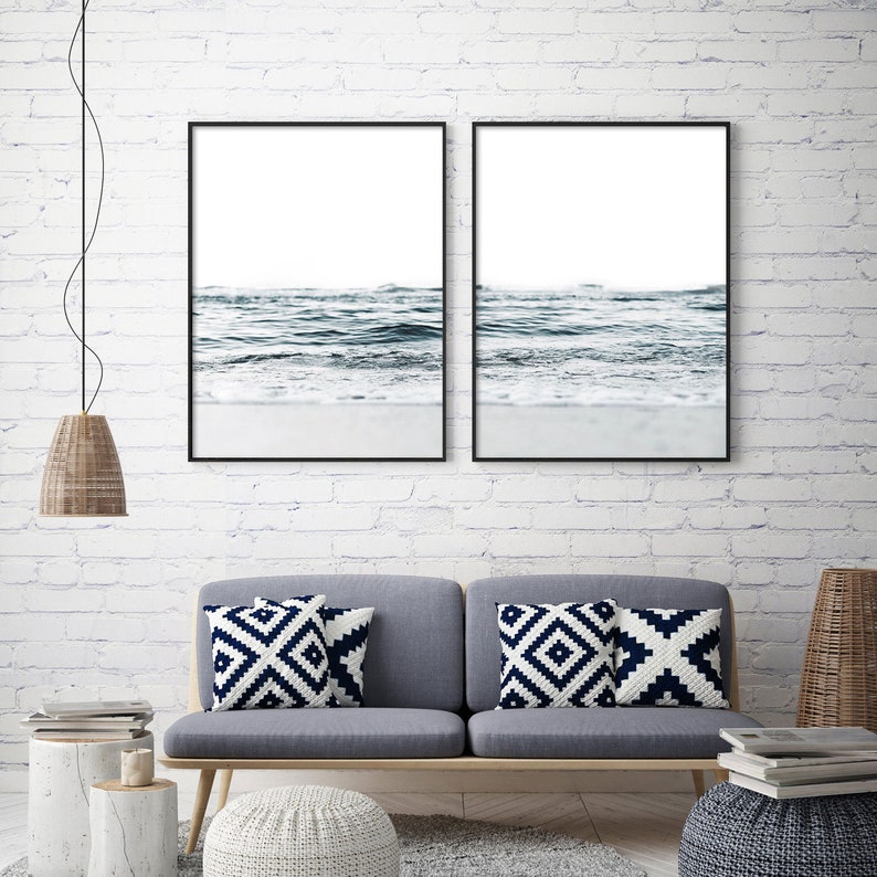 May include: Two framed black and white photographs of ocean waves crashing on a sandy beach. The photos are hung on a white brick wall above a gray couch with white and blue patterned throw pillows.