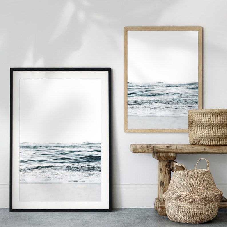 May include: Two framed photographs of ocean waves with a white sandy beach in the foreground. The photographs are hanging on a white wall above a wooden bench with two woven baskets.