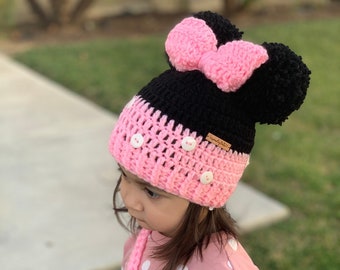 May include: A pink and black crocheted hat with a bow on top and two pom-poms. The hat has white buttons on the front. The hat is being worn by a child wearing a pink and white polka dot shirt.