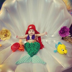 May include: A baby dressed as a mermaid sits in a white inflatable seashell. The baby is wearing a red crochet hat with a white star, a purple bikini top, and a green crochet mermaid tail. There are yellow and orange crocheted sea creatures and a yellow plush fish on the seashell.