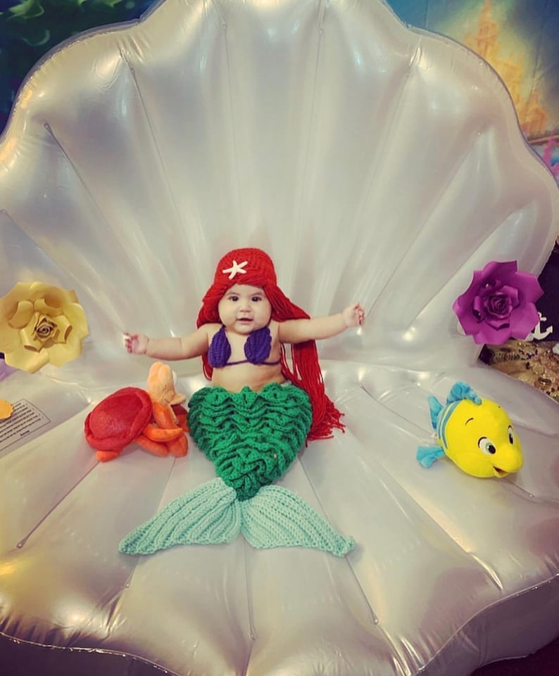 May include: A baby dressed as a mermaid sits in a white inflatable seashell. The baby is wearing a red crochet hat with a white star, a purple bikini top, and a green crochet mermaid tail. There are yellow and orange crocheted sea creatures and a yellow plush fish on the seashell.