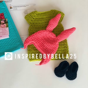 May include: A crocheted outfit for a baby, including a green dress, a pink bunny hat, and black shoes. The outfit is handmade and features a delicate crocheted texture.
