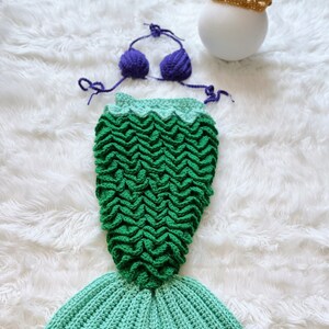 May include: A green and blue crocheted mermaid tail with a purple crocheted bikini top and a gold crocheted crown.