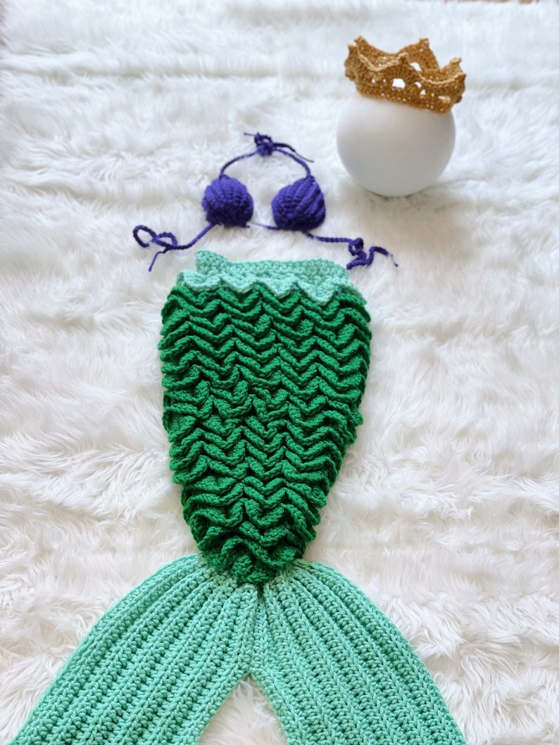May include: A green and blue crocheted mermaid tail with a purple crocheted bikini top and a gold crocheted crown.