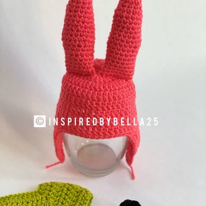 May include: A pink crocheted bunny hat with long floppy ears. The hat has a brim and a long flap that can be tied under the chin. The hat is sitting on a glass surface. The image also includes a green crocheted shirt and two black crocheted shoes.