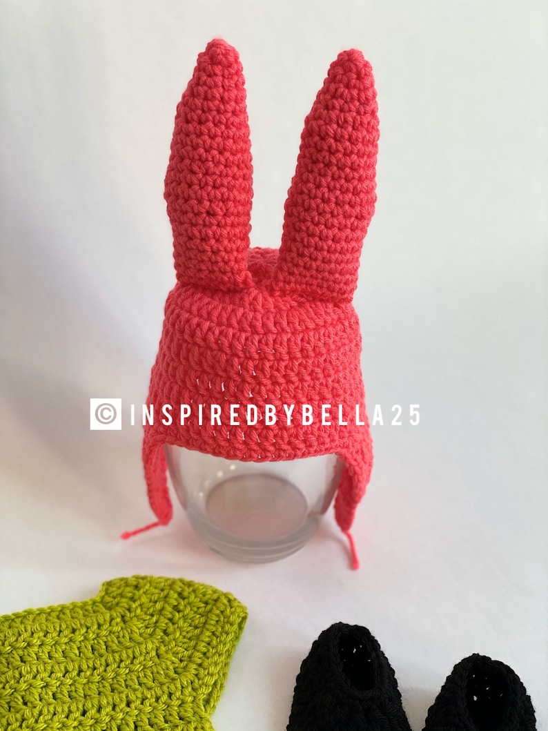 May include: A pink crocheted bunny hat with long floppy ears. The hat has a brim and a long flap that can be tied under the chin. The hat is sitting on a glass surface. The image also includes a green crocheted shirt and two black crocheted shoes.