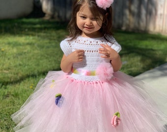 May include: A young girl wearing a white crocheted top with a pink tulle skirt decorated with crocheted flowers. She is wearing a pink headband with a flower.