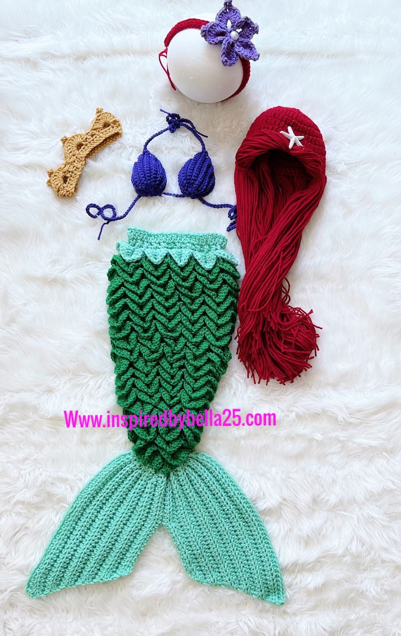 May include: A crocheted mermaid costume set for a baby, featuring a green and blue tail, a purple flower headband, a red wig with a white star, and a blue bikini top.