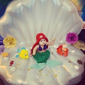 May include: A baby dressed as a mermaid sits in a large white inflatable seashell. The baby is wearing a red crochet hat with a white star, a green crochet mermaid tail, and a purple crochet top. The seashell is decorated with gold glitter, white seashells, and other nautical decorations.