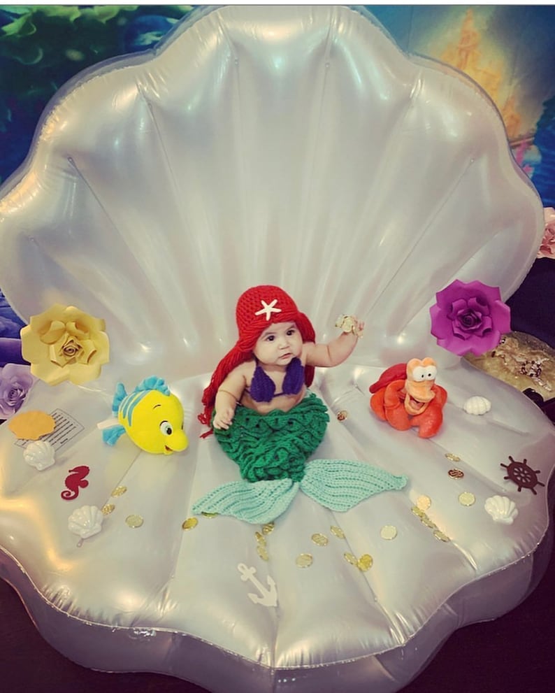 May include: A baby dressed as a mermaid sits in a large white inflatable seashell. The baby is wearing a red crochet hat with a white star, a green crochet mermaid tail, and a purple crochet top. The seashell is decorated with gold glitter, white seashells, and other nautical decorations.