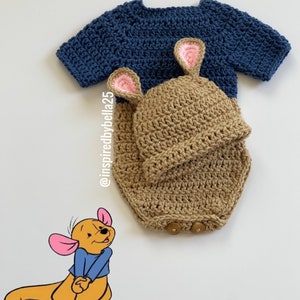 May include: A crocheted baby outfit in brown and blue with a kangaroo design. The outfit includes a hat with ears and a onesie with buttons. The outfit is inspired by Winnie the Pooh's character, Roo.