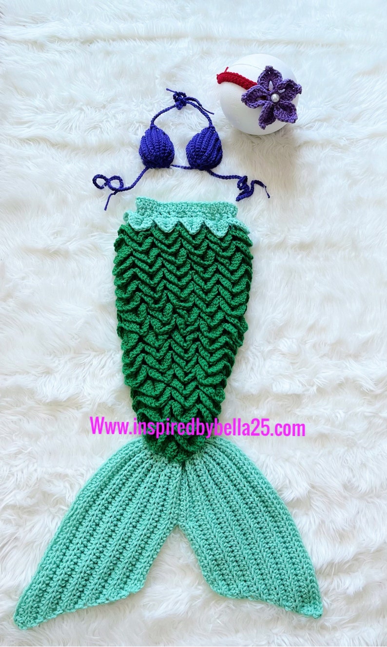 May include: A green and blue crocheted mermaid tail costume with a purple crocheted bikini top and a purple flower headband. The costume is on a white fluffy surface. www.inspiredbybella25.com