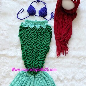 May include: A crocheted mermaid tail costume for a baby or toddler. The tail is green and turquoise with a scale pattern. The set also includes a purple crochet bikini top and a red crocheted wig with a white starfish.