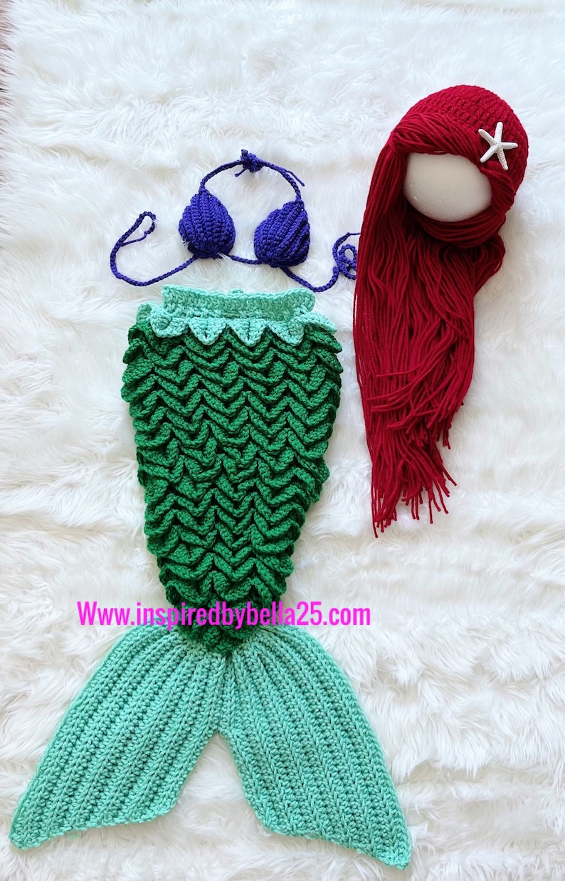 May include: A crocheted mermaid tail costume for a baby or toddler. The tail is green and turquoise with a scale pattern. The set also includes a purple crochet bikini top and a red crocheted wig with a white starfish.