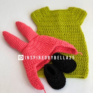 May include: A crocheted baby outfit set in green, pink, and black. The set includes a dress, a hat with bunny ears, and a pair of booties.