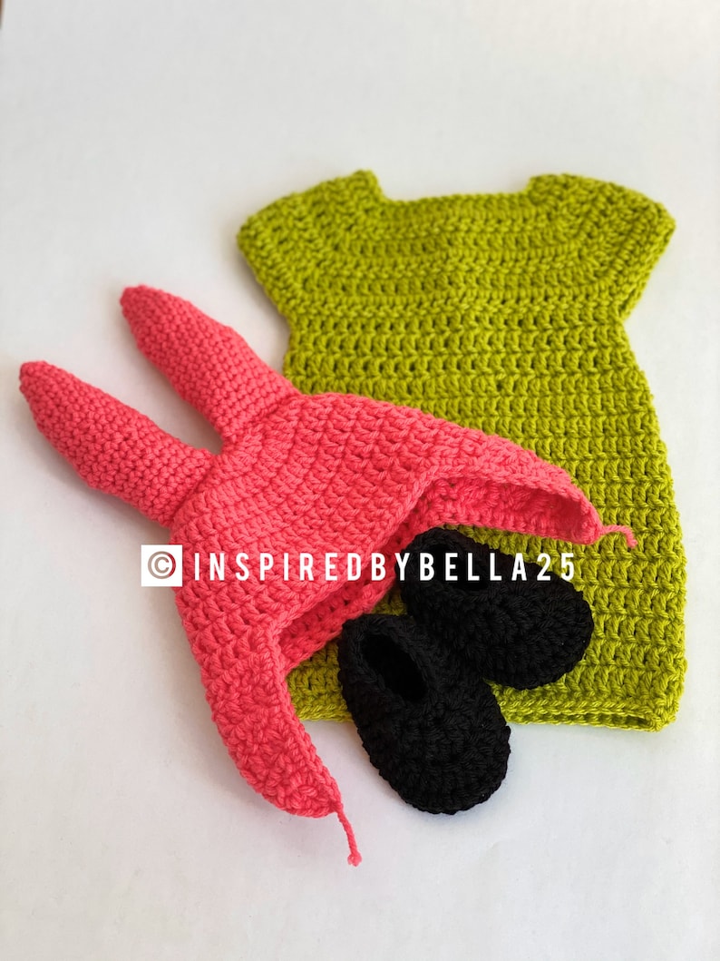 May include: A crocheted baby outfit set in green, pink, and black. The set includes a dress, a hat with bunny ears, and a pair of booties.