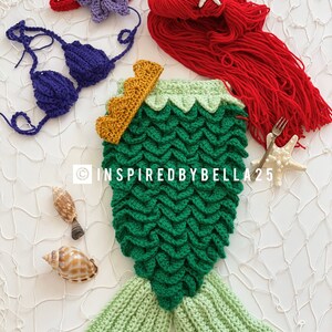 May include: A crocheted mermaid tail costume for a baby, featuring green scales, a light green tail fin, and a gold crown. The costume is laid out on a white background with seashells and a starfish.
