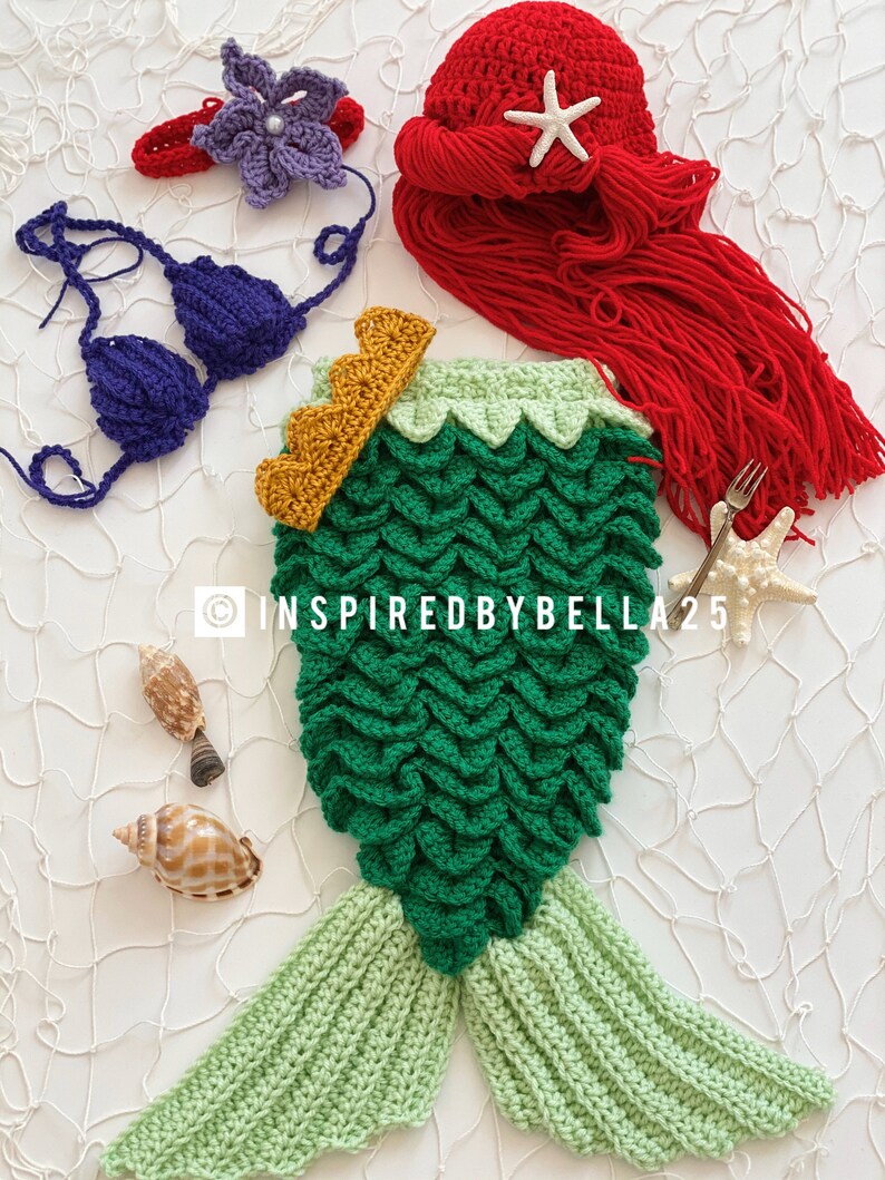May include: A crocheted mermaid tail costume for a baby, featuring green scales, a light green tail fin, and a gold crown. The costume is laid out on a white background with seashells and a starfish.