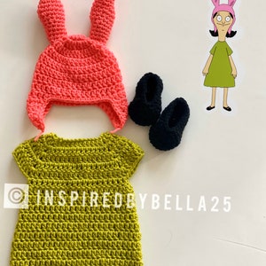 May include: A crocheted green dress with short sleeves, a pink crocheted bunny hat with long ears, and two black crocheted shoes. The image includes the text "INSPIRED BY BELLA 25".