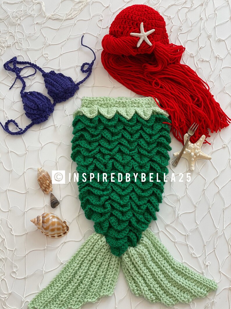 May include: A green crocheted mermaid tail with a light green tail fin. A purple crocheted bikini top and a red crocheted hat with a white starfish are also included. The image is on a white background with a white fishing net.