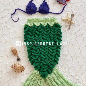 May include: A crocheted mermaid tail costume for a baby, featuring a green tail with a light green ombre effect, a green and light green top, and a purple crochet bikini top. The costume is laid out on a white background with seashells and a starfish.