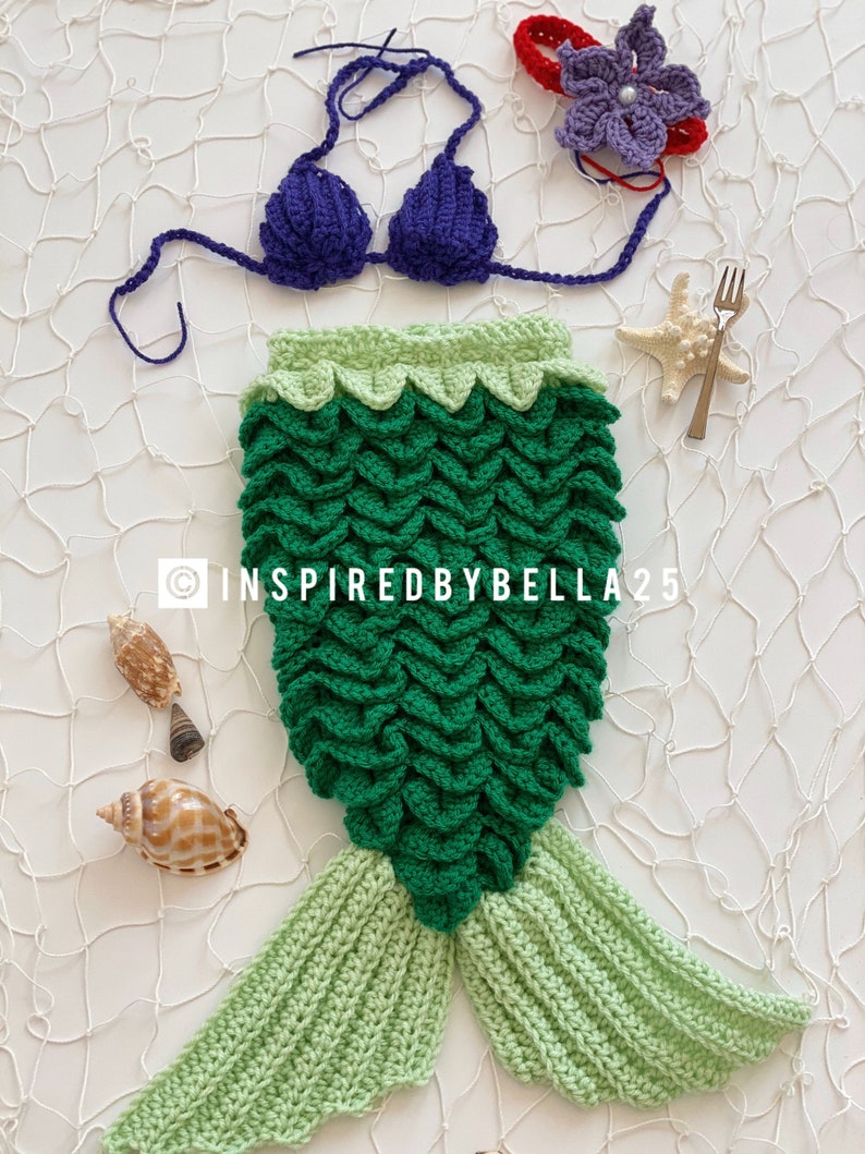 May include: A crocheted mermaid tail costume for a baby, featuring a green tail with a light green ombre effect, a green and light green top, and a purple crochet bikini top. The costume is laid out on a white background with seashells and a starfish.
