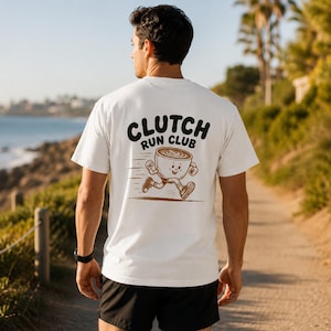 以下が含まれることがあります： 背面に茶色のグラフィックが施された白い半袖Tシャツ。デザインには、手足が付いて走っている漫画風のコーヒーカップが描かれており、イラストの上部には太字の黒いテキストでCLUTCH RUN CLUBと記されています。