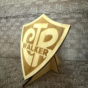 May include: A wooden shield-shaped sign with the letters "CTP" and the name "WALKER" engraved in a light brown color. The sign is propped up by a wooden stand. The background is a textured, gray surface.