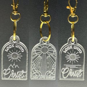 May include: Three clear acrylic keychains with gold metal rings and clips. Each keychain features a sun, clouds, and the text "Look Unto Christ".