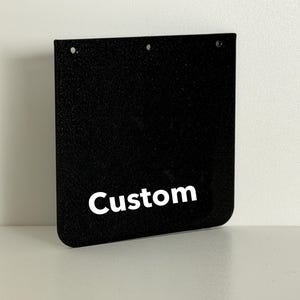 Puede incluir: Un guardabarros negro con superficie texturizada. La palabra "Custom" está impresa en letras blancas en la parte inferior. Se ven tres agujeros en la parte superior para el montaje.