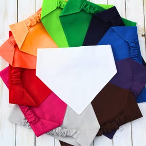 May include: A selection of colourful pet bandanas in shades of orange, green, blue, and purple. Each bandana is a different colour and has an elastic band. The bandanas are arranged in a circular pattern on a white wooden surface.
