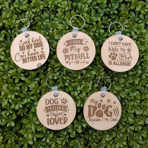 May include: Five wooden keychains with different dog-themed sayings. The keychains are round and have a brown wood finish. The sayings include "I work hard so my dog can have a better life", "I love my pitbull", "I can't have kids my dog is allergic", "Dog mother coffee lover", and "My dog thinks I'm cool".