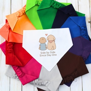 May include: A collection of colorful dog bandanas and matching headbands. The central bandana is white with a cartoon illustration of a baby and a dog, and the text "Side by Side Since Day One". The bandanas and headbands are in various colors.