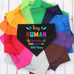 以下が含まれることがあります： 赤、ピンク、オレンジ、緑など、カラフルなペット用バンダナのコレクション。中央の黒いバンダナには、虹色の「Tiny HUMAN Security」の文字と、手形と矢印のグラフィックが描かれています。バンダナは円形に配置されています。