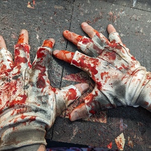 May include: Two white fingerless gloves with red blood stains on them. The gloves are lying on a dark gray surface.