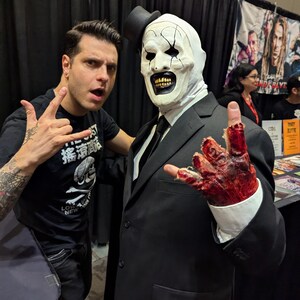 May include: A man in a black suit and a white clown mask with a black top hat is standing next to a man with tattoos on his arm. The man in the suit has a bloody hand and is making a rock and roll hand gesture.