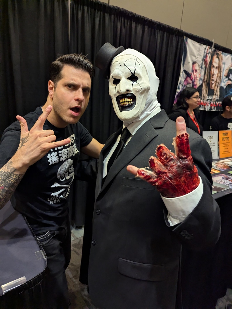 May include: A man in a black suit and a white clown mask with a black top hat is standing next to a man with tattoos on his arm. The man in the suit has a bloody hand and is making a rock and roll hand gesture.