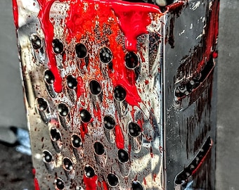 May include: A stainless steel box grater with a red liquid substance covering the surface. The grater has a handle on the top and is covered in red liquid.