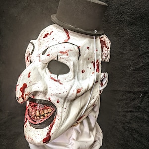 May include: A white creepy clown mask with a black top hat. The mask has a wide, exaggerated smile with visible teeth and is covered in red splatters of paint that resemble blood.