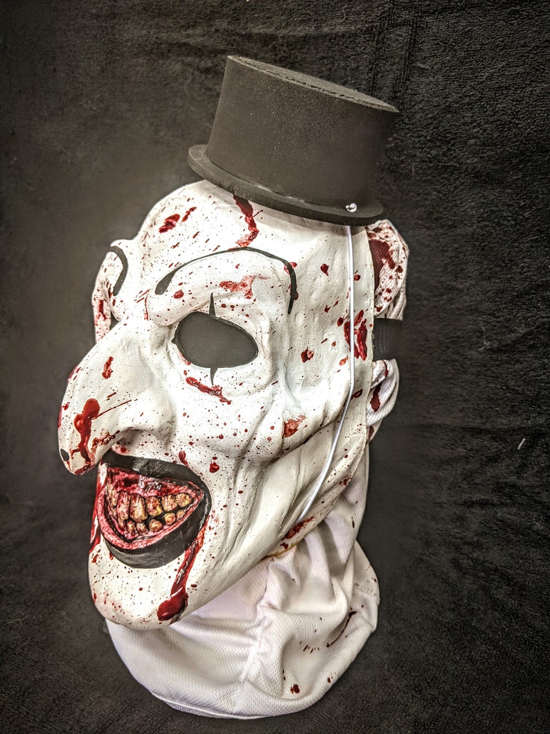 May include: A white creepy clown mask with a black top hat. The mask has a wide, exaggerated smile with visible teeth and is covered in red splatters of paint that resemble blood.