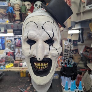 May include: A white creepy clown mask with black painted lines and a black top hat. The mask has a wide, toothy grin and black eyes.