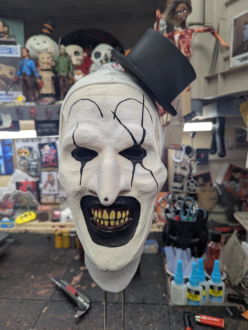 May include: A white creepy clown mask with black painted lines and a black top hat. The mask has a wide, toothy grin and black eyes.