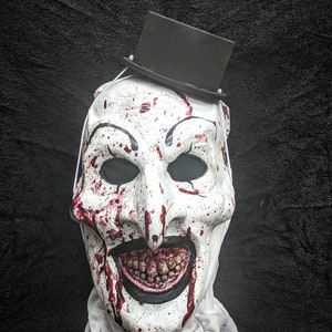 May include: A white creepy clown mask with a black top hat, splattered with red paint, and a wide, toothy grin.
