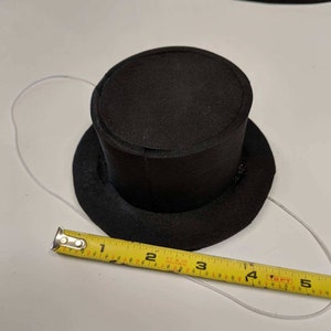 May include: A black foam top hat with a white string attached to the brim. A yellow measuring tape is placed below the hat, showing measurements in inches.