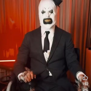 May include: A person wearing a white mask with black eyes and a black mouth, a black suit, and a black top hat, sitting in a wheelchair. The person is holding a silver object in their right hand. The background is a red curtain.