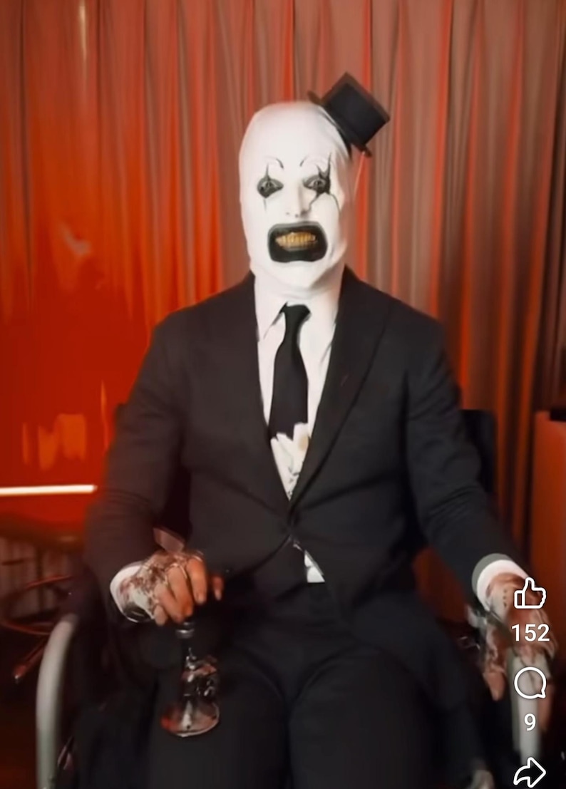 May include: A person wearing a white mask with black eyes and a black mouth, a black suit, and a black top hat, sitting in a wheelchair. The person is holding a silver object in their right hand. The background is a red curtain.
