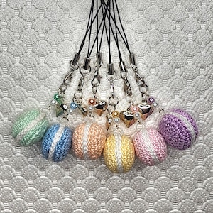 May include: A set of seven colorful crocheted macaron charms with silver heart accents and pearl beads. Each charm is attached to a black string with a gold metal clasp.
