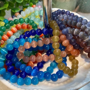 Pode incluir: Uma coleção de pulseiras coloridas feitas com contas redondas e iridescentes. As pulseiras estão dispostas em círculo sobre um prato branco.