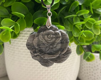 May include: A gray and black stone rose pendant necklace with a silver bail. The pendant is carved in the shape of a rose and has a detailed design. The necklace is on a black cord.