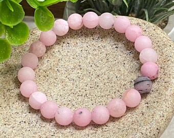 May include: A pink gemstone bracelet with a matte finish. The bracelet is made of round beads and has a dark grey bead.